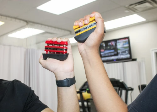Two people raise small hand grip strengtheners for a grip workout in a gym, one red model and one yellow/black model visible in the foreground.