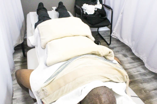 Person lying face down on a massage table, covered with towels and pillows in a treatment room.
