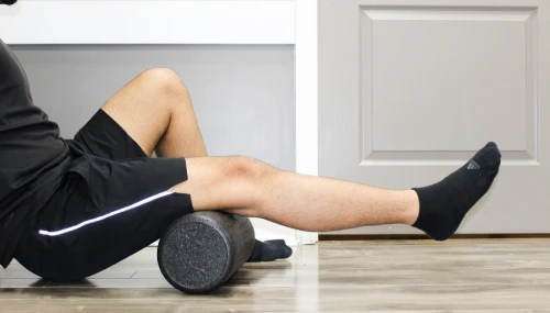 Person lying on a wooden floor, using a foam roller under their calves with one leg extended, wearing black shorts and socks.