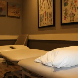 Treatment room with a padded massage/exam table, a white pillow at the foot, and framed wall art above.