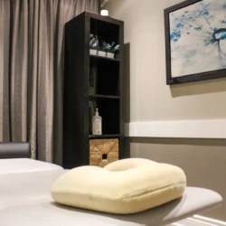 Spa treatment room with a white massage table and a beige neck pillow, framed by dark curtains and a tall black shelf.