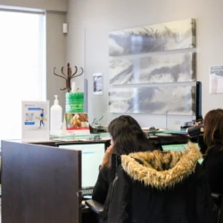 Two people sit at a reception desk in an office, facing computer screens with coats draped nearby.