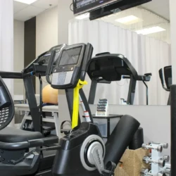 Indoor gym scene featuring a black stationary bike in the foreground, with a treadmill behind it, a weight rack to the right, and a large mirror on the back wall. A ceiling monitor is mounted above.