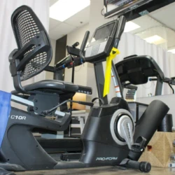 Recumbent exercise bike with a digital display in a bright gym space; a blue strap hangs from the side.