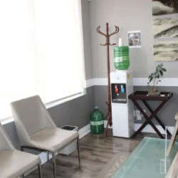 Small waiting area with beige chairs, a glass coffee table, and a water dispenser against a gray wall.