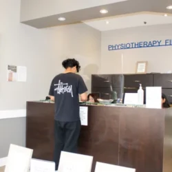 Reception at a physiotherapy clinic with a dark wood desk; a man stands at the counter while staff assist him; 'PHYSIOTHERAPY FIRST' sign on the wall.